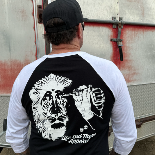 A person wearing the Not All There 3/4 Sleeve Raglan T Shirt by Life Out There Apparel stands with their back to the camera, displaying a lion’s face being tattooed by a hand holding a tattoo machine.