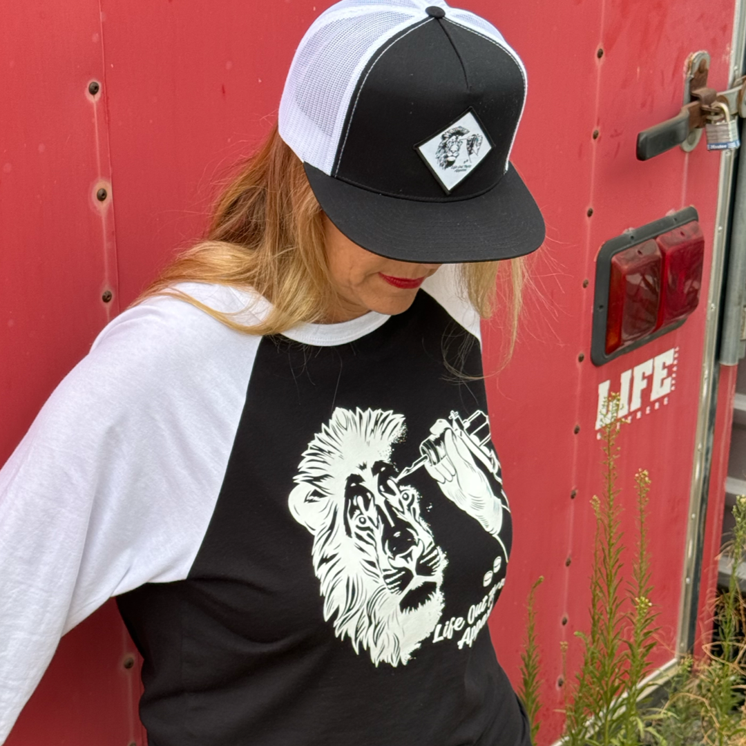 A woman in the Not All There 3/4 Sleeve Raglan T Shirt by Life Out There Apparel leans against a red wall with a LIFE sign, glancing down—an image promoting suicide prevention. Black, white, and lion graphic; green plants nearby.