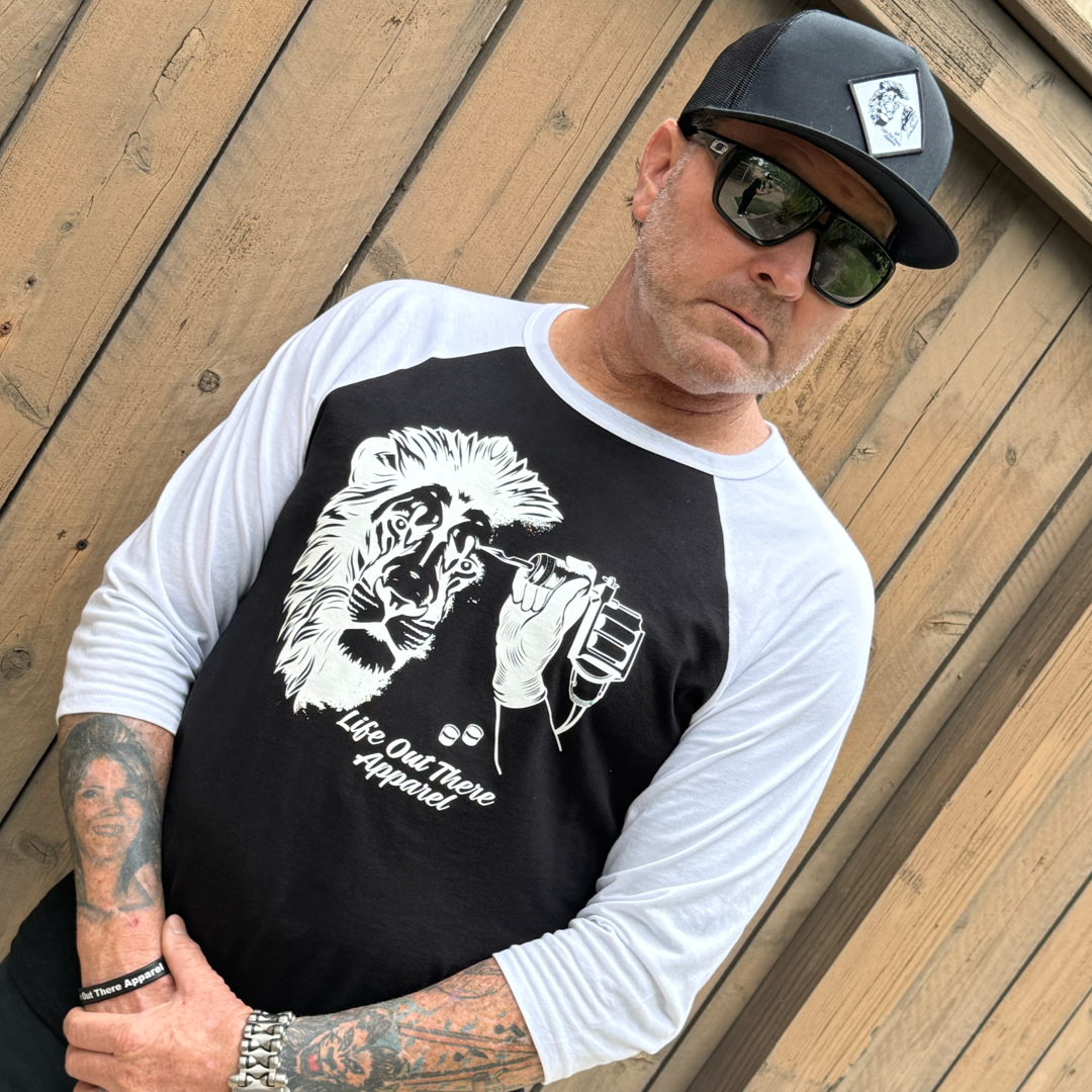 A man in Life Out There Apparel’s Unisex Not All There 3/4 Sleeve Raglan T Shirt (Black/White) stands before a wooden fence, wearing sunglasses and a black cap, showing off tattooed arms as he clasps his hands.