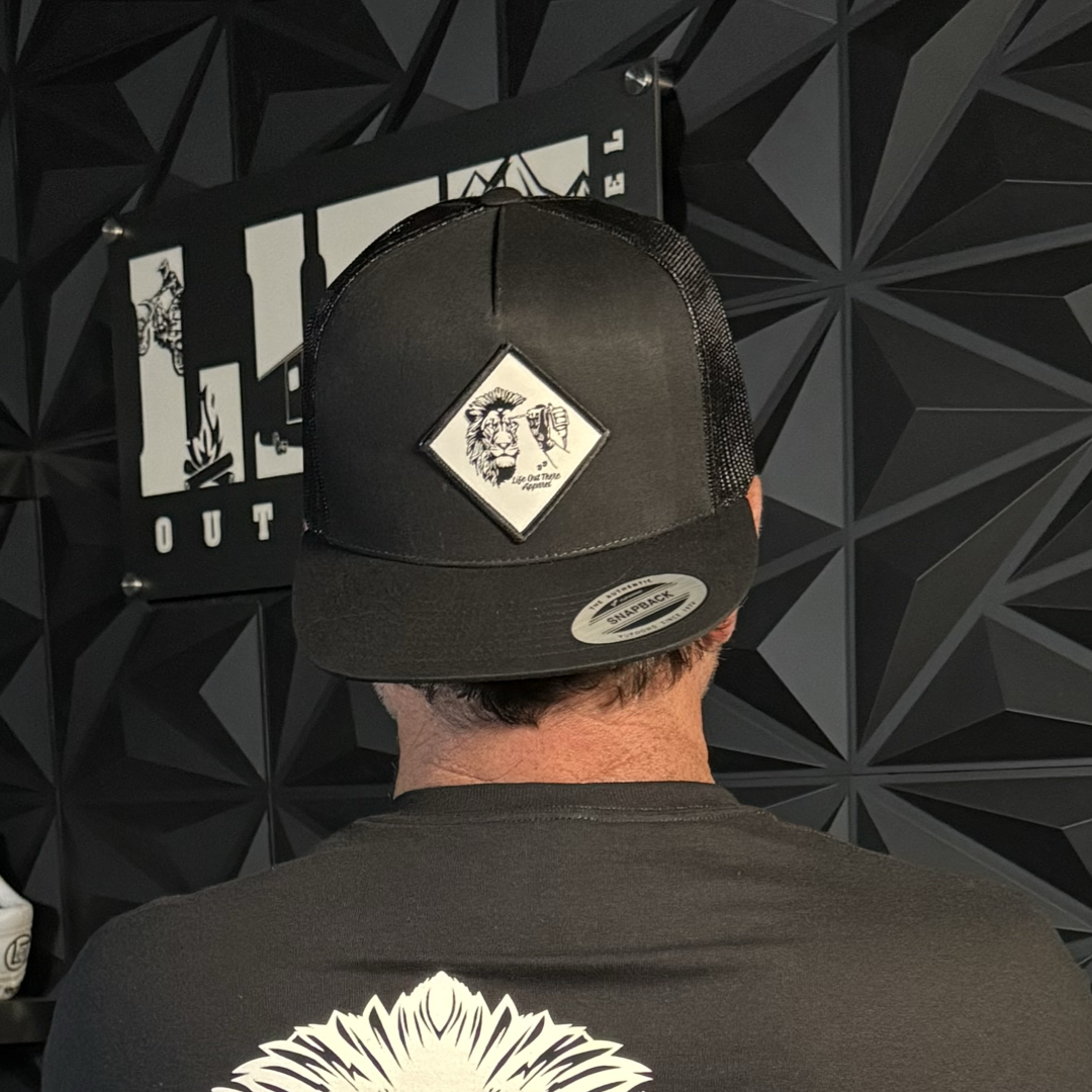 A person wears the Not All There - Flat Bill SnapBack Trucker Hat in black by Life Out There Apparel, featuring a diamond-shaped animal patch, while standing before a textured black wall with a partially visible sign.