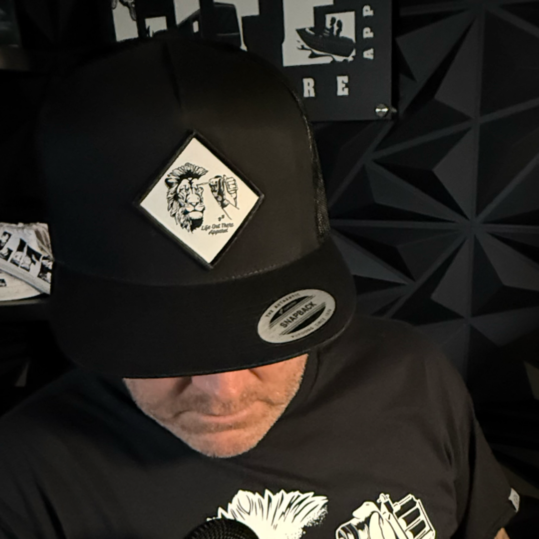 A person in a Life Out There Apparel Not All There - Flat Bill SnapBack Trucker Hat in black looks down, wearing a matching black shirt. The background displays geometric patterns and part of a sign with bold white letters.