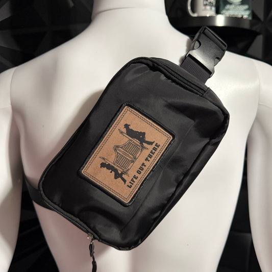 The Yeehaw— a medium black fanny pack/hand bag by Life Out There Apparel—features zip pockets and a tan patch with two hikers and “LIFE OUT THERE,” shown on a white mannequin torso.