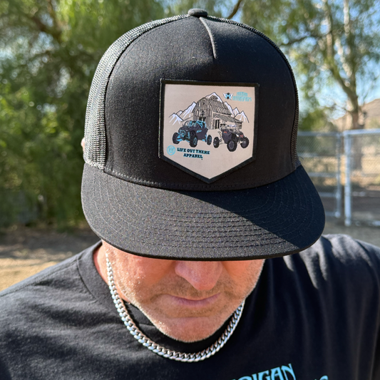 A person wears the Life Out There Apparel Side X Side - Flat Bill SnapBack Trucker Hat in black, featuring a patch with mountains and an off-road vehicle. They also have on a silver chain necklace and black shirt, with trees and a fence in the background.