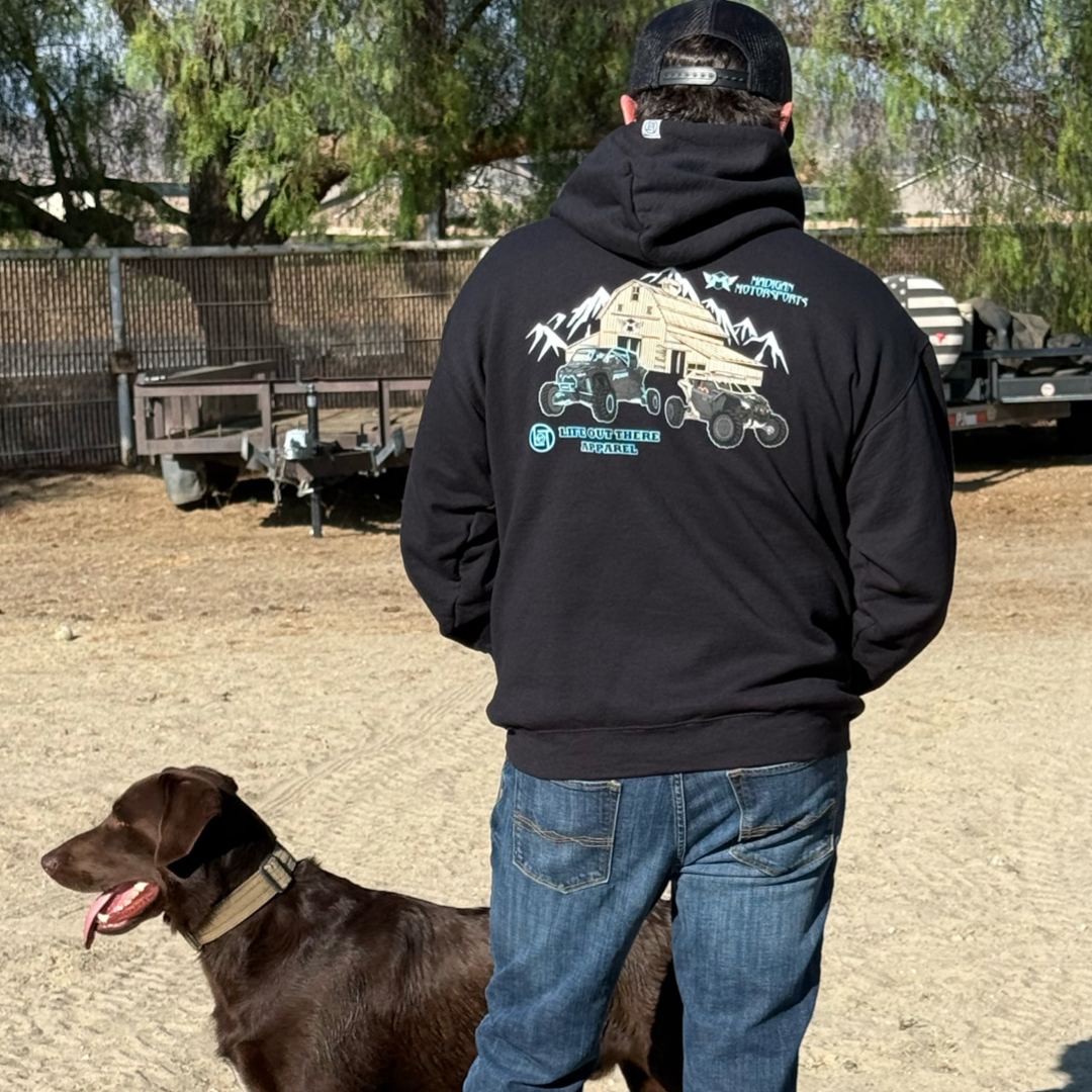 A man in a Life Out There Apparel Unisex Side X Side Hoodie (black) stands on dirt beside a brown dog, wearing off-road apparel. Trailers, trees, and a fence are visible outdoors at Madigan Motorsports.
