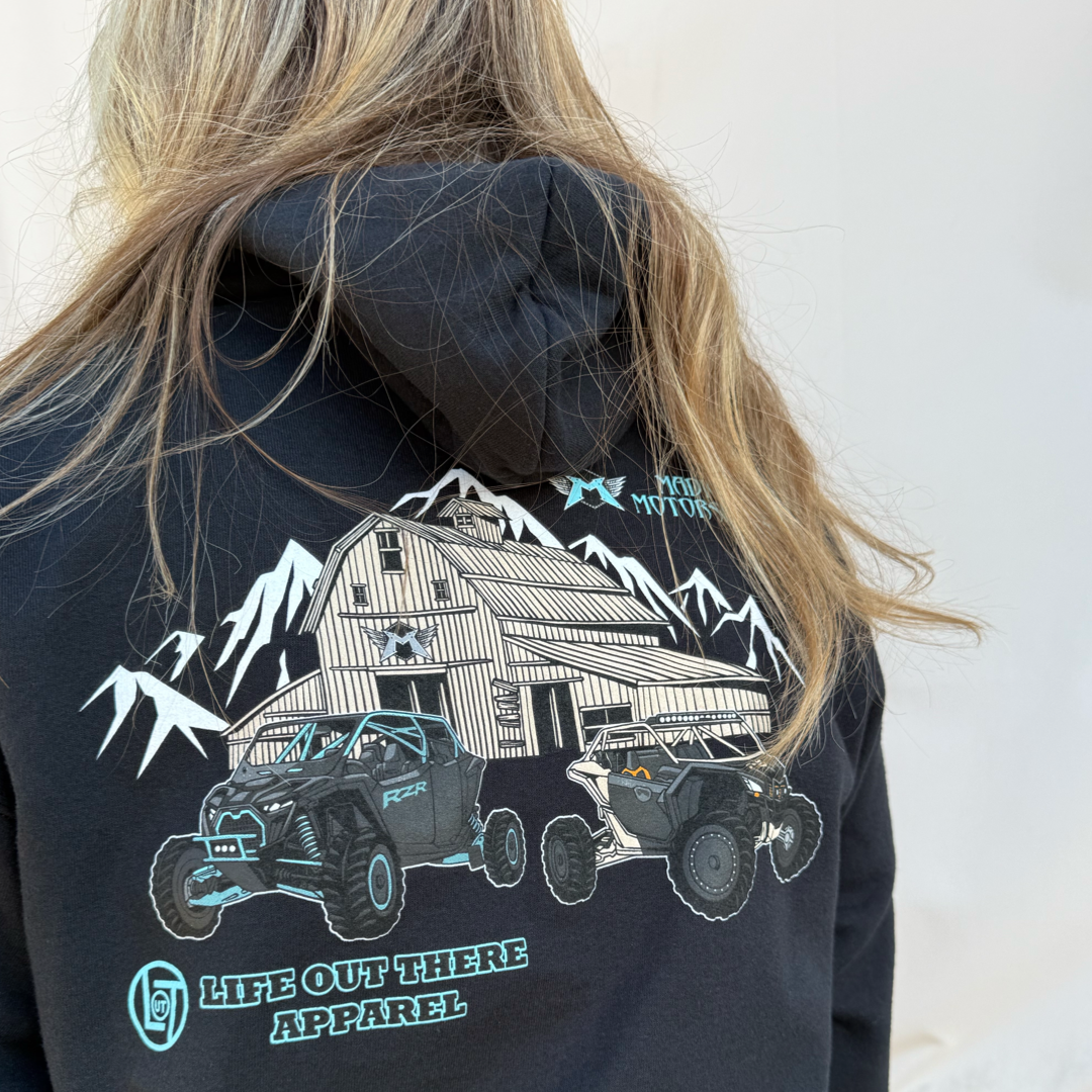A person with long hair wears the Unisex - Side X Side Hoodie in black by Life Out There Apparel, featuring a barn, mountains, and two Side X Side vehicles—ideal off-road gear inspired by Madigan Motorsports.