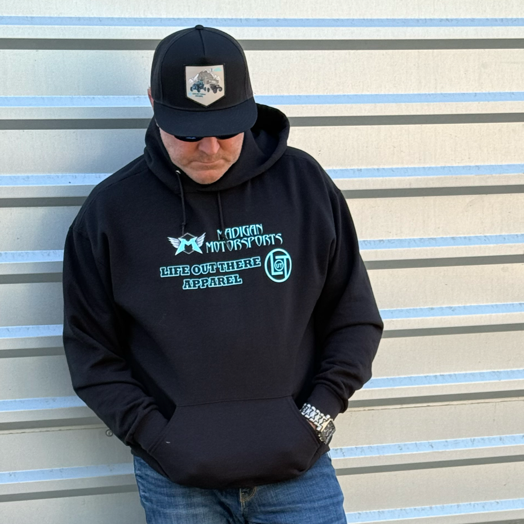 A man in sunglasses, a black "Unisex - Side X Side Hoodie - Black" by Life Out There Apparel, a black cap, and a wristwatch stands with hands in his pockets before a striped metal wall.