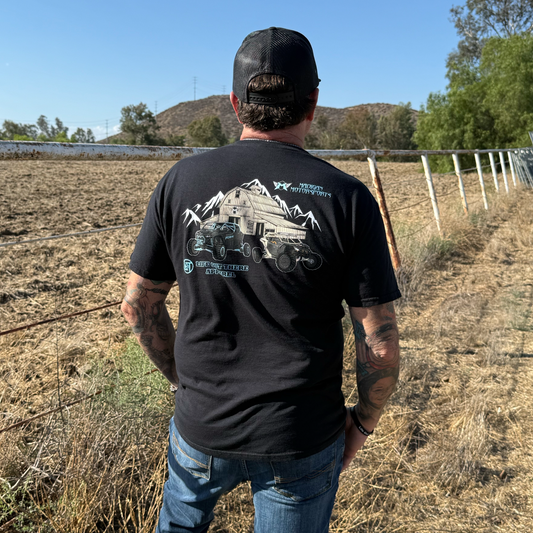 A man wearing the Life Out There Apparel Side X Side Tee - Black stands by a dry, fenced field with hills and trees, dressed in blue jeans and a black cap. His tattooed arms and graphic tee capture an adventurous, off-road spirit.