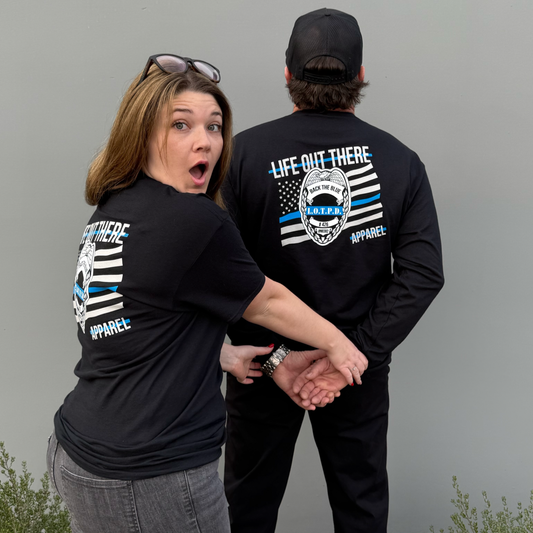 A surprised woman pretends to handcuff a man from behind. Both wear Life Out There Apparel's "Back The Blue" long sleeve black tees, showing support for law enforcement against a plain gray wall.