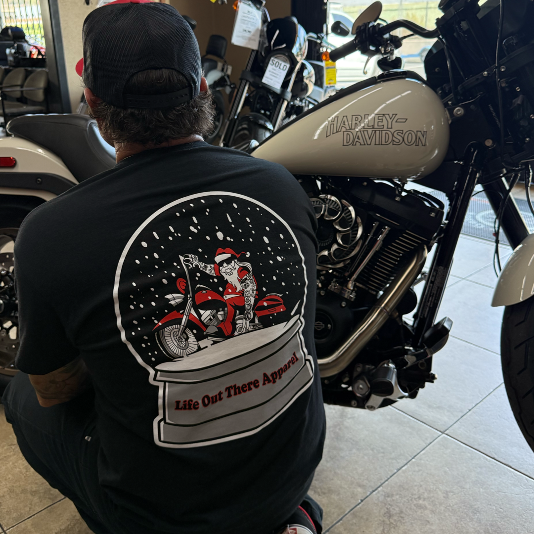 A person in the Life Out There Apparel Unisex - Dashing Through The Snow Tee (Black), featuring Santa on a motorcycle, sits beside a silver Harley-Davidson in a showroom.