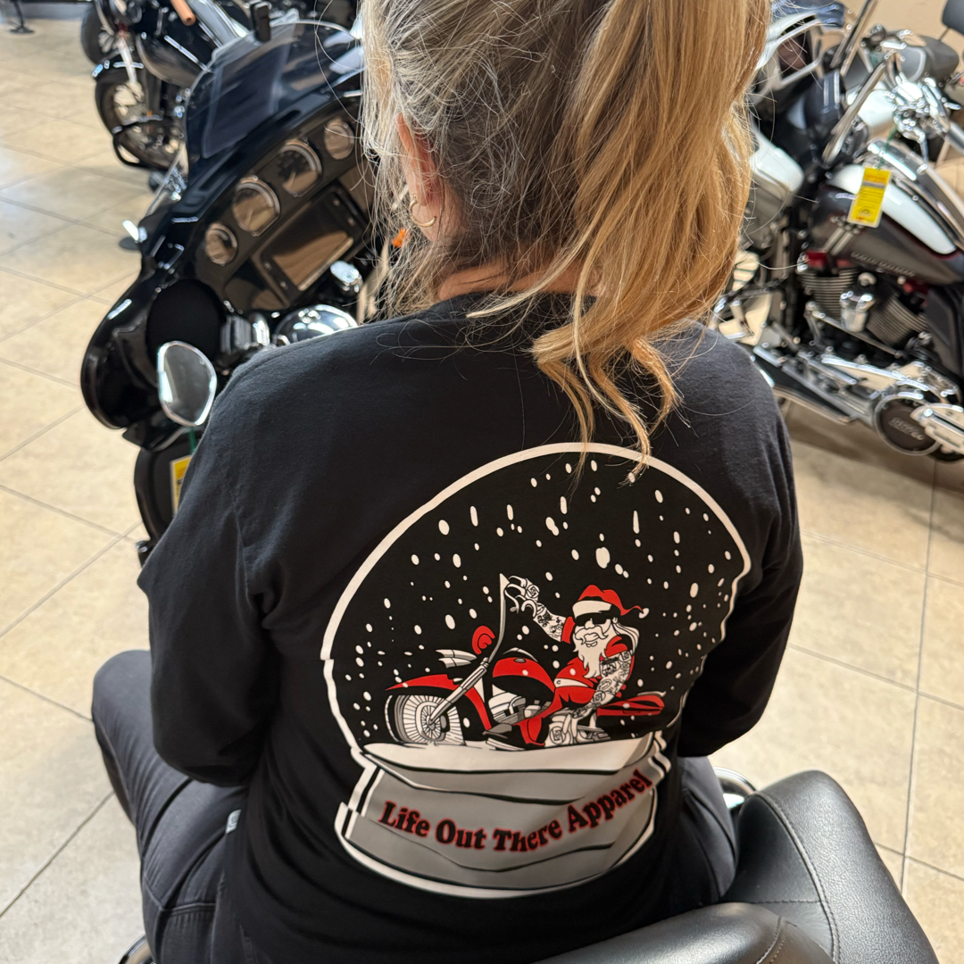 A blonde woman sits indoors on a motorcycle, wearing Life Out There Apparel's Unisex Dashing Through The Snow Tee in black, featuring cartoon Santa Claus on a red bike. Other motorcycles are visible in the background.