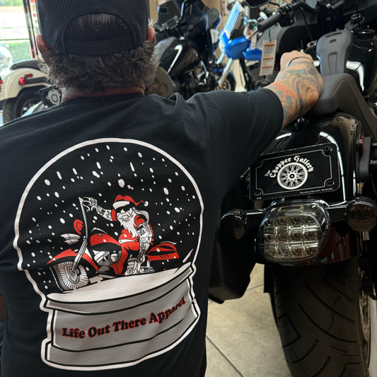 A bearded man in a cap sits on a black motorcycle, wearing the Dashing Through The Snow Tee from Life Out There Apparel, which features Santa on a red bike in a snow globe with snowfall and the brand's name at the base.