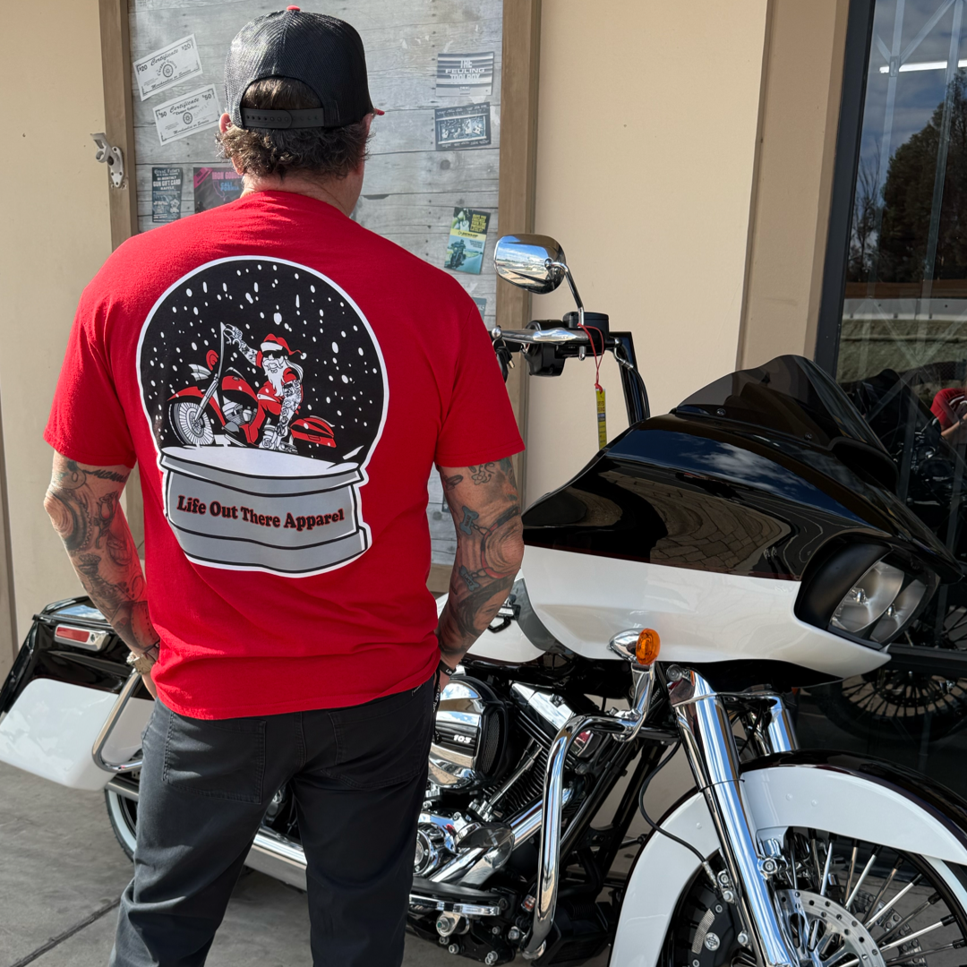 A man in a Life Out There Apparel Unisex "Dashing Through The Snow" red tee stands beside a black and white motorcycle, facing away from the camera on a sunny day.