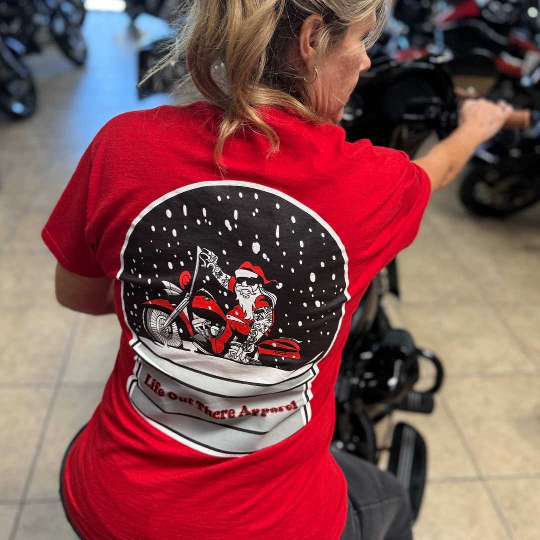 A woman wears the limited edition "Dashing Through The Snow" red tee by Life Out There Apparel, featuring a snow globe with Santa riding a motorcycle, as she sits on a motorcycle indoors.