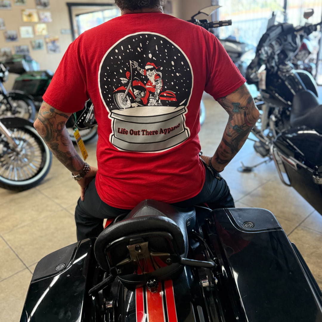 A person wearing the limited edition "Unisex - Dashing Through The Snow Tee - Red" from Life Out There Apparel, featuring a snow globe motorcycle graphic, sits on a black motorcycle inside a showroom with other bikes in the background.