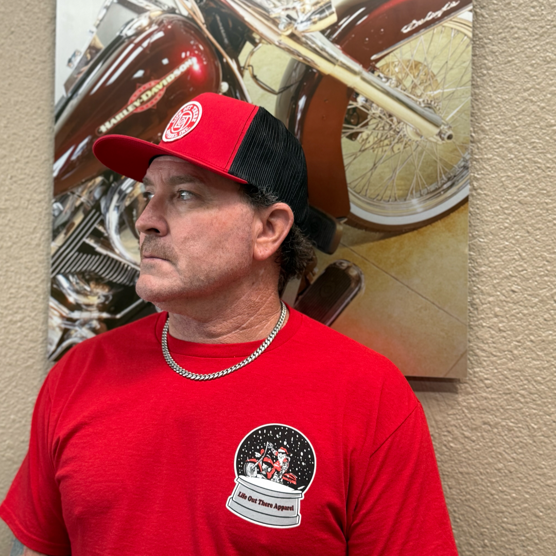 A man in a red Life Out There Apparel Unisex - Dashing Through The Snow Tee and a red-and-black cap stands before a motorcycle painting, looking left and wearing a silver chain; the limited edition tee features a graphic logo on the chest.