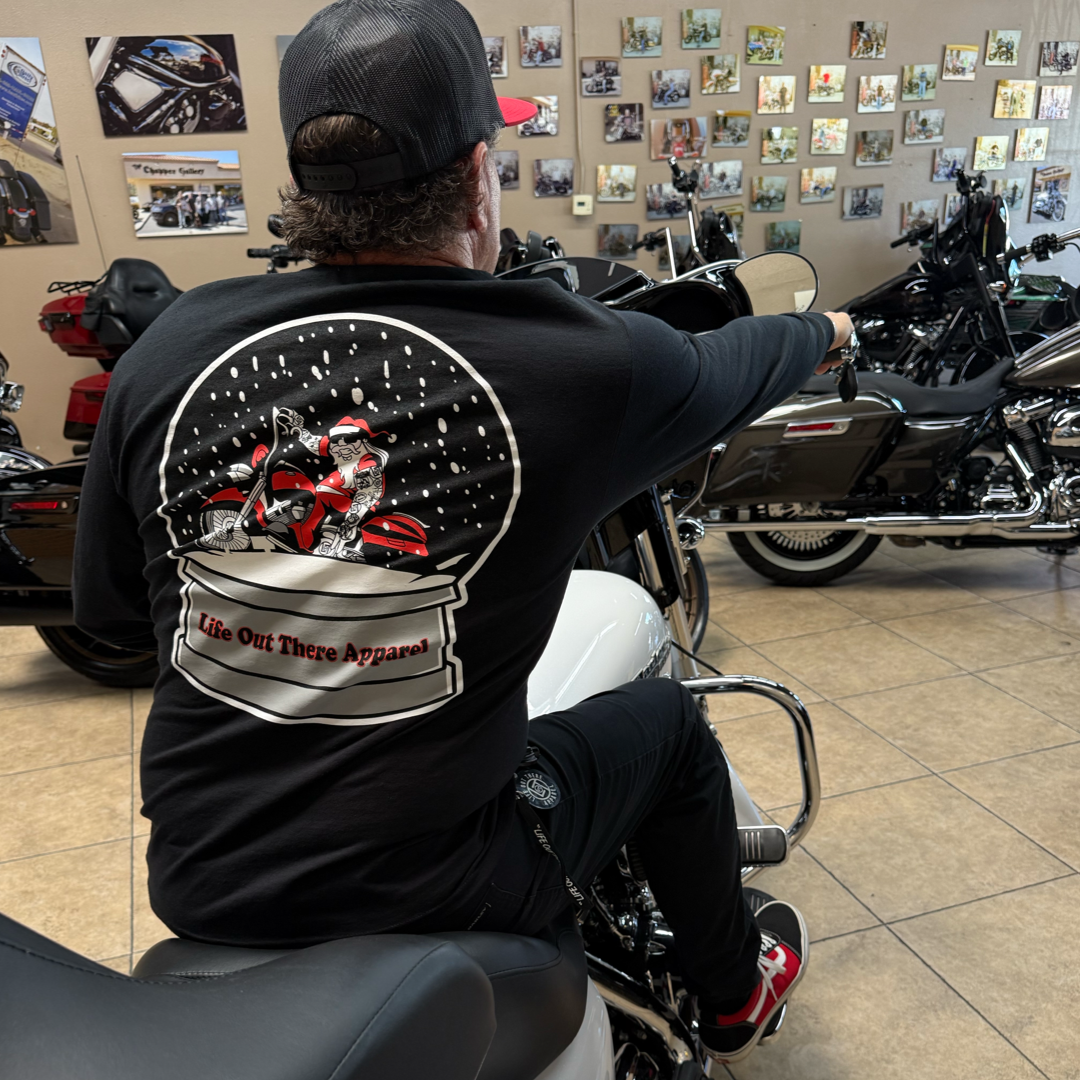 A man in a black cap and the Unisex - Dashing Through The Snow - Long Sleeve Tee by Life Out There Apparel, featuring a motorcycle snow globe design, sits on a white motorcycle in a showroom filled with bikes and wall photos.