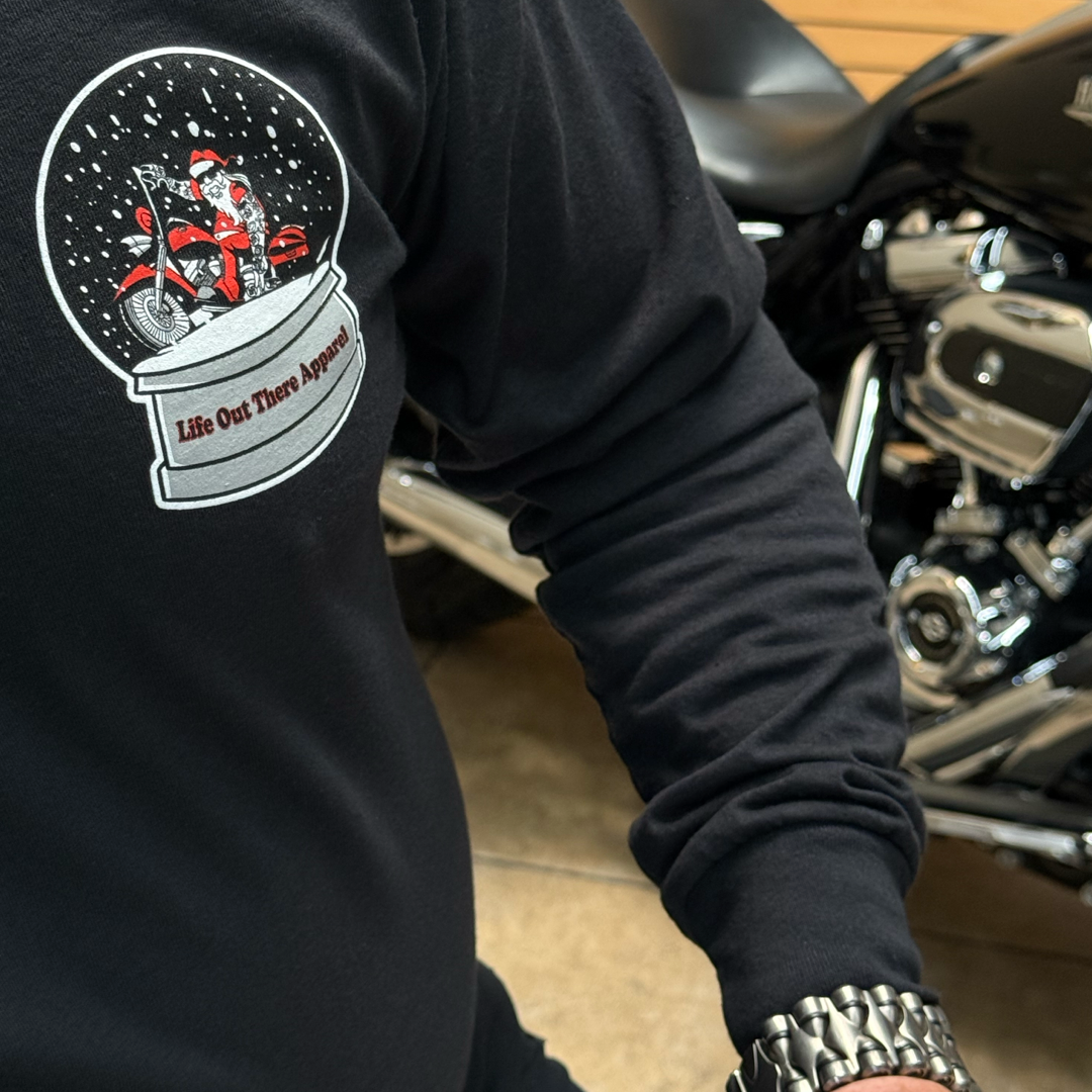 A person in the “Unisex - Dashing Through The Snow - Long Sleeve Tee” by Life Out There Apparel, featuring Santa riding a motorcycle in the snow, stands next to a parked bike. This black limited edition tee is perfect for festive riders.