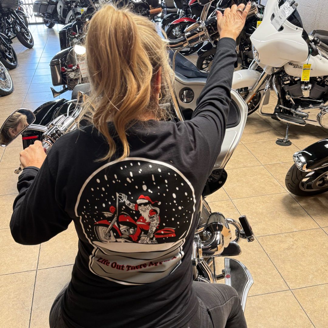 A blonde woman in a ponytail sits on a motorcycle in a showroom, wearing Life Out There Apparel’s Unisex - Dashing Through The Snow - Long Sleeve Tee in black, featuring Santa riding a red motorcycle.