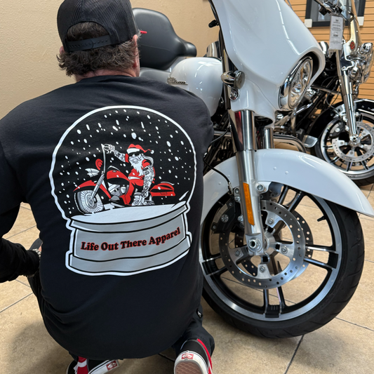 Wearing a black cap and the "Dashing Through The Snow - Long Sleeve Tee" by Life Out There Apparel, featuring Santa on a motorcycle snow globe, a person sits indoors beside a white motorcycle.