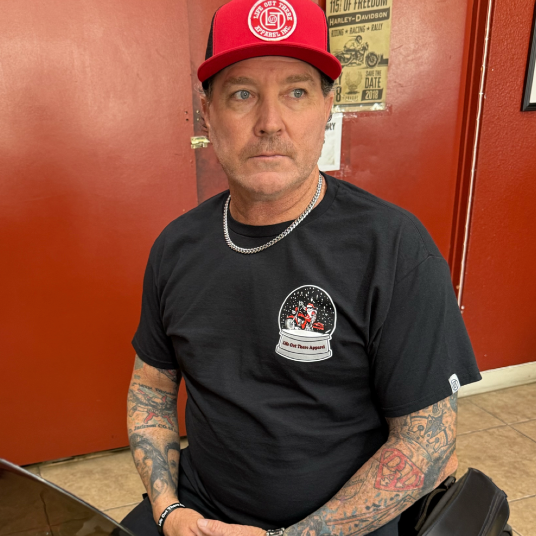 A tattooed man sits indoors, wearing a black "Unisex - Dashing Through The Snow Tee" by Life Out There Apparel and a red cap. He looks serious, seated before red walls decorated with posters.
