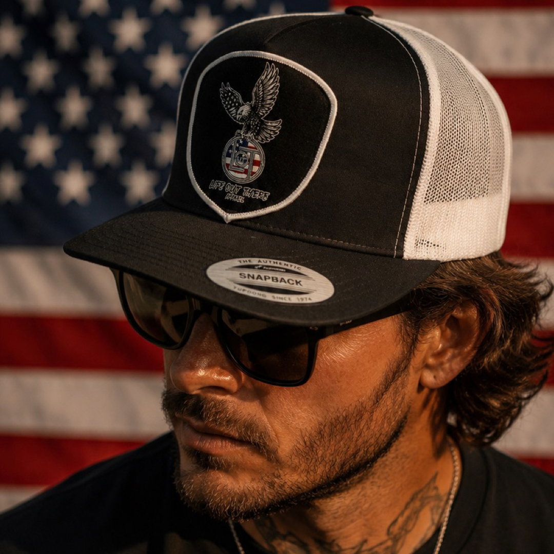 A man with wavy brown hair, a beard, and sunglasses wears the Life Out There Apparel Flight of Freedom Flat Bill SnapBack Trucker Hat (Black/White) while standing before a large, blurred American flag.
