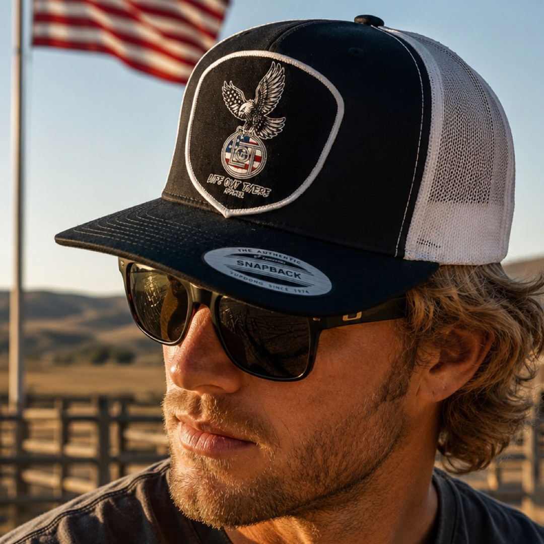 A man in sunglasses wears the Life Out There Apparel Flight of Freedom Flat Bill SnapBack Trucker Hat (Black/White) featuring an eagle emblem, standing outdoors with an American flag and sunlit landscape behind him.