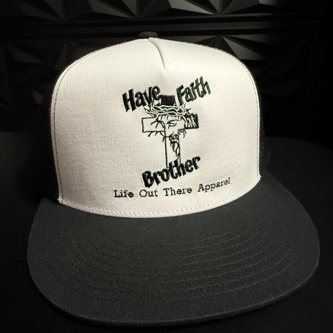 The Have Faith Brother (Embroidered) Flat Bill SnapBack Trucker Hat in White/Black by Life Out There Apparel features bold text above and below a crowned cross, with the brand name embroidered beneath. The hat is shown on a dark surface.