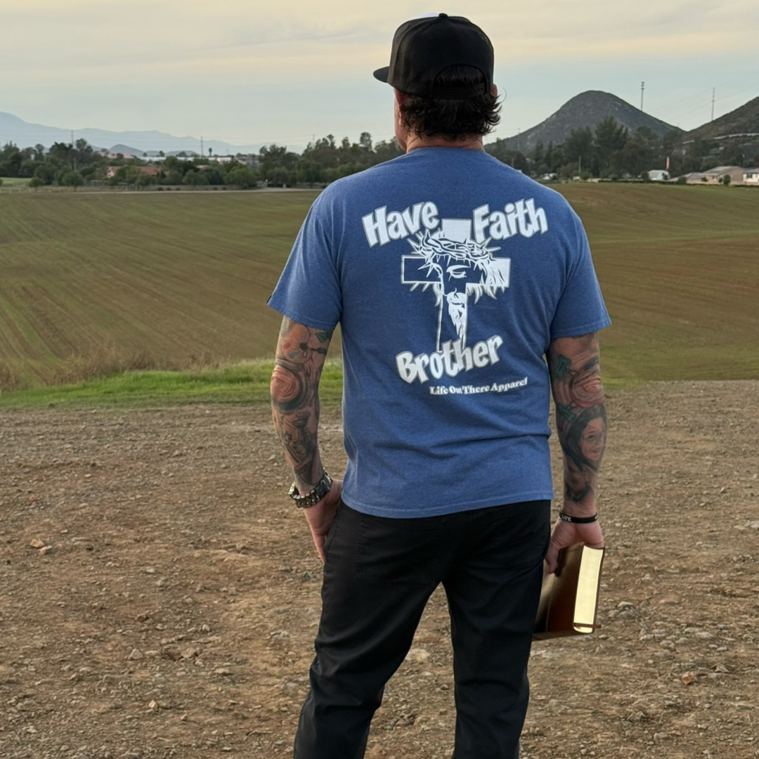 A tattooed man in a black cap stands outdoors on a dirt path, holding a book and wearing the Unisex - Have Faith Brother Tee in Vintage Blue from Life Out There Apparel, facing a field with hills and houses in the distance.