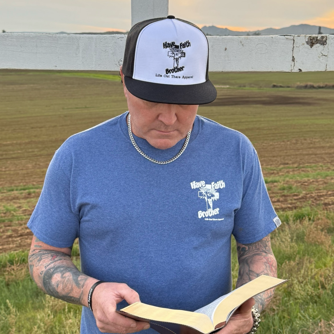 A tattooed man in a vintage blue "Have Faith Brother" tee by Life Out There Apparel stands outside before a field and wooden cross, reading a book—capturing the spirit of Christian faithwear.
