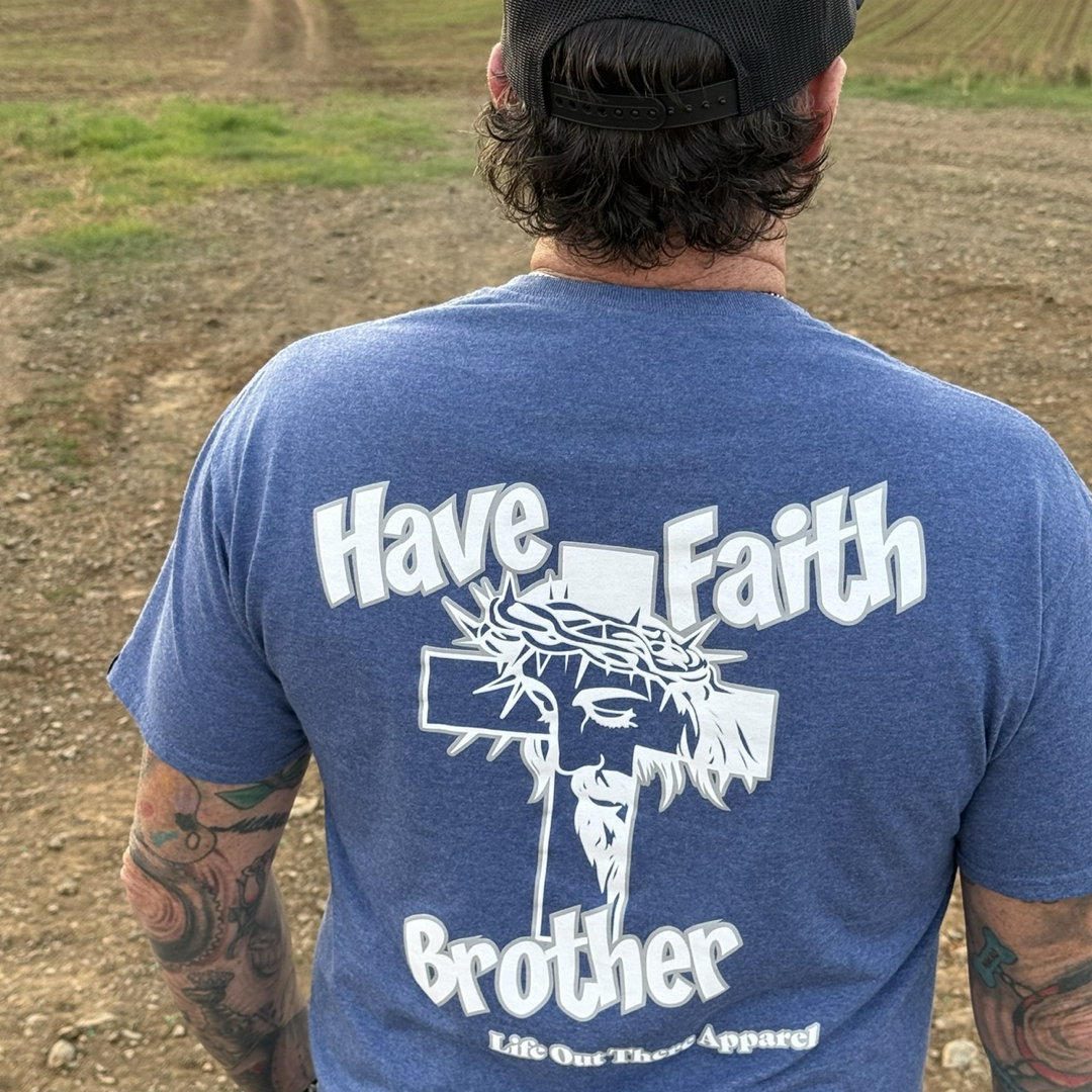 Wearing the Have Faith Brother Tee in Vintage Blue by Life Out There Apparel, a tattooed person faces away, showcasing the bold Christian message: "HAVE FAITH BROTHER" alongside a cross and Jesus' face.