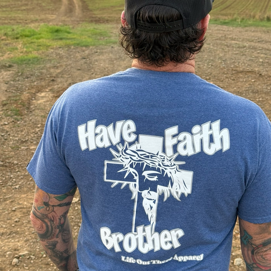 A person stands outdoors on a dirt path wearing the Unisex - Have Faith Brother Tee in Vintage Blue by Life Out There Apparel, featuring "HAVE FAITH BROTHER" with Jesus, a crown of thorns, and a cross. Their back and tattooed arms are visible.