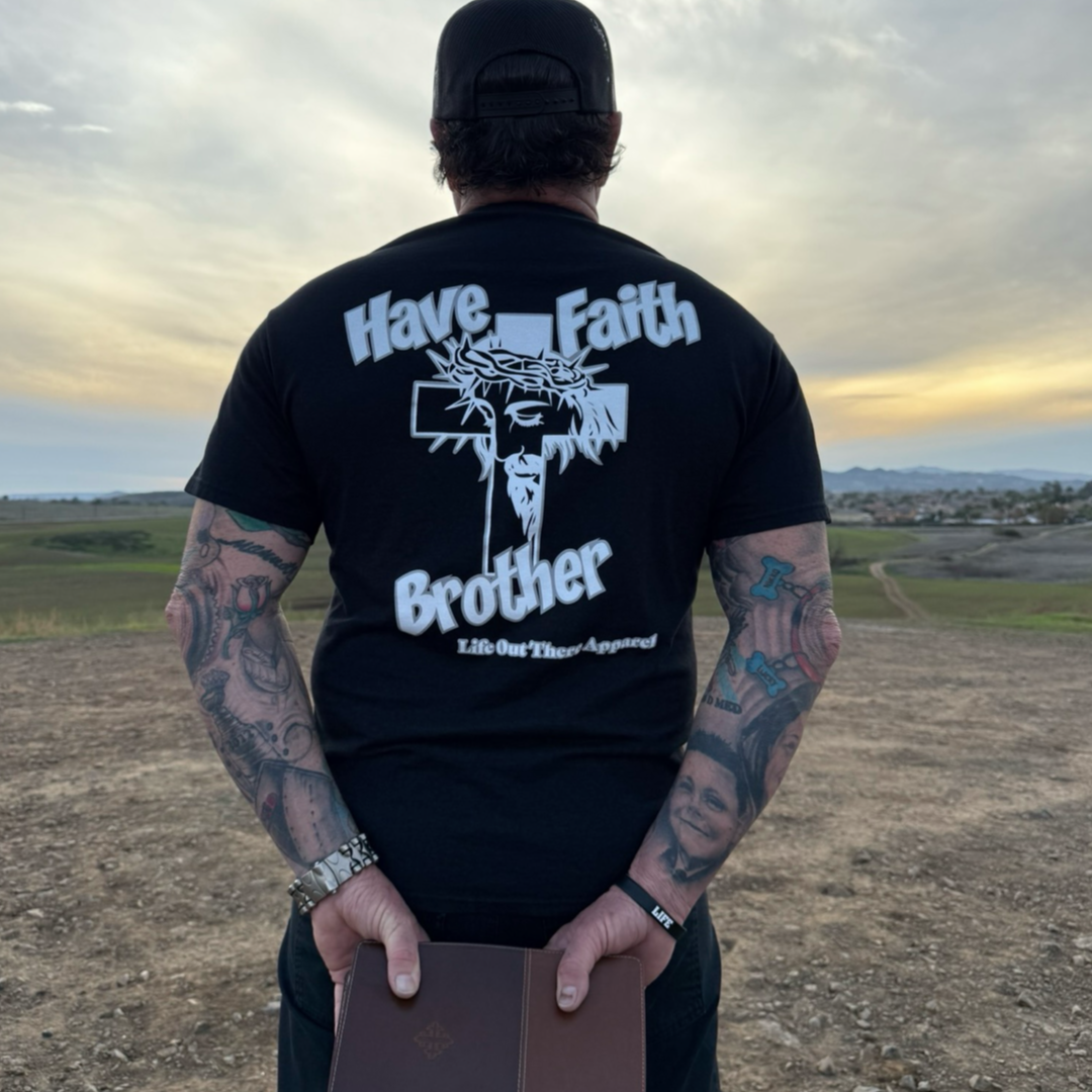 A tattooed man stands outdoors at sunset, facing away. He holds a Bible behind his back and wears the black “Have Faith Brother” unisex tee by Life Out There Apparel, featuring a cross and crown of thorns.