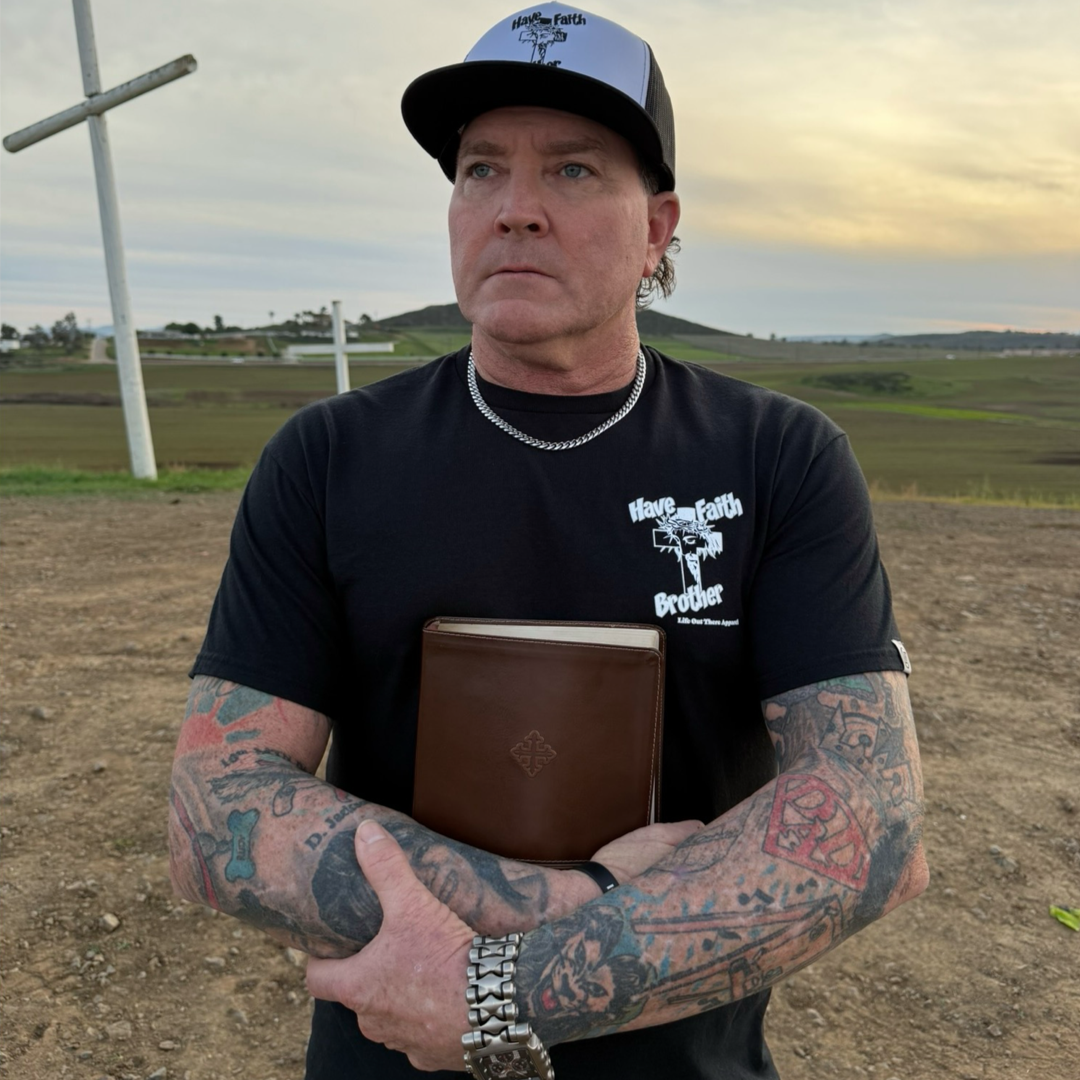 A tattooed man in a black "Have Faith Brother" tee by Life Out There Apparel and a baseball cap stands outdoors, holding a closed Bible near large white crosses, with a sunset landscape behind him.