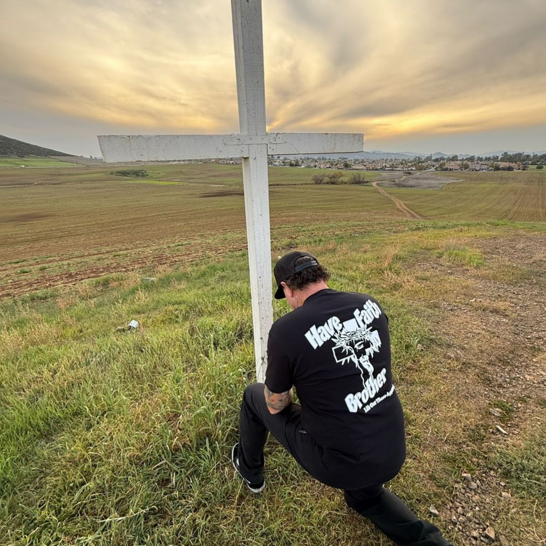 A man wearing the Unisex - Have Faith Brother Tee in black by Life Out There Apparel kneels beside a white cross on a grassy hill at sunset, with cloudy skies and distant fields visible.