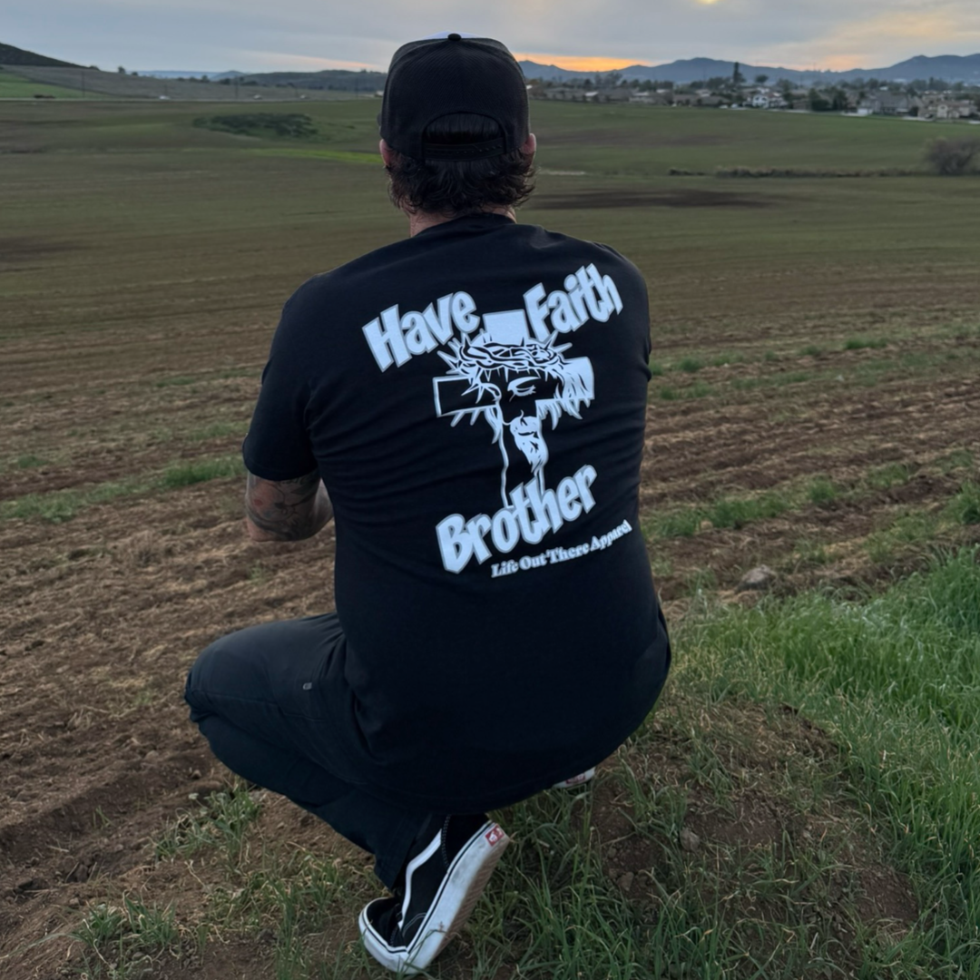 Kneeling on grass in black attire and a cap, a person overlooks farmland while wearing the Life Out There Apparel Unisex - Have Faith Brother Tee in Black, featuring "HAVE FAITH BROTHER" with a cross and crown graphic.