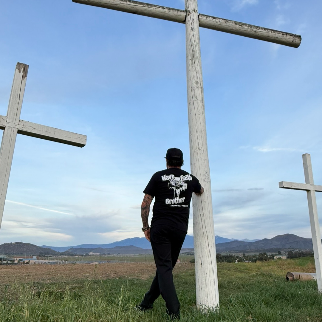 A person in black stands among large white crosses outdoors, resting a hand on a cross and gazing at distant mountains beneath a blue sky. They wear the Life Out There Apparel Unisex - Have Faith Brother Tee - Black, expressing their Christian faith.