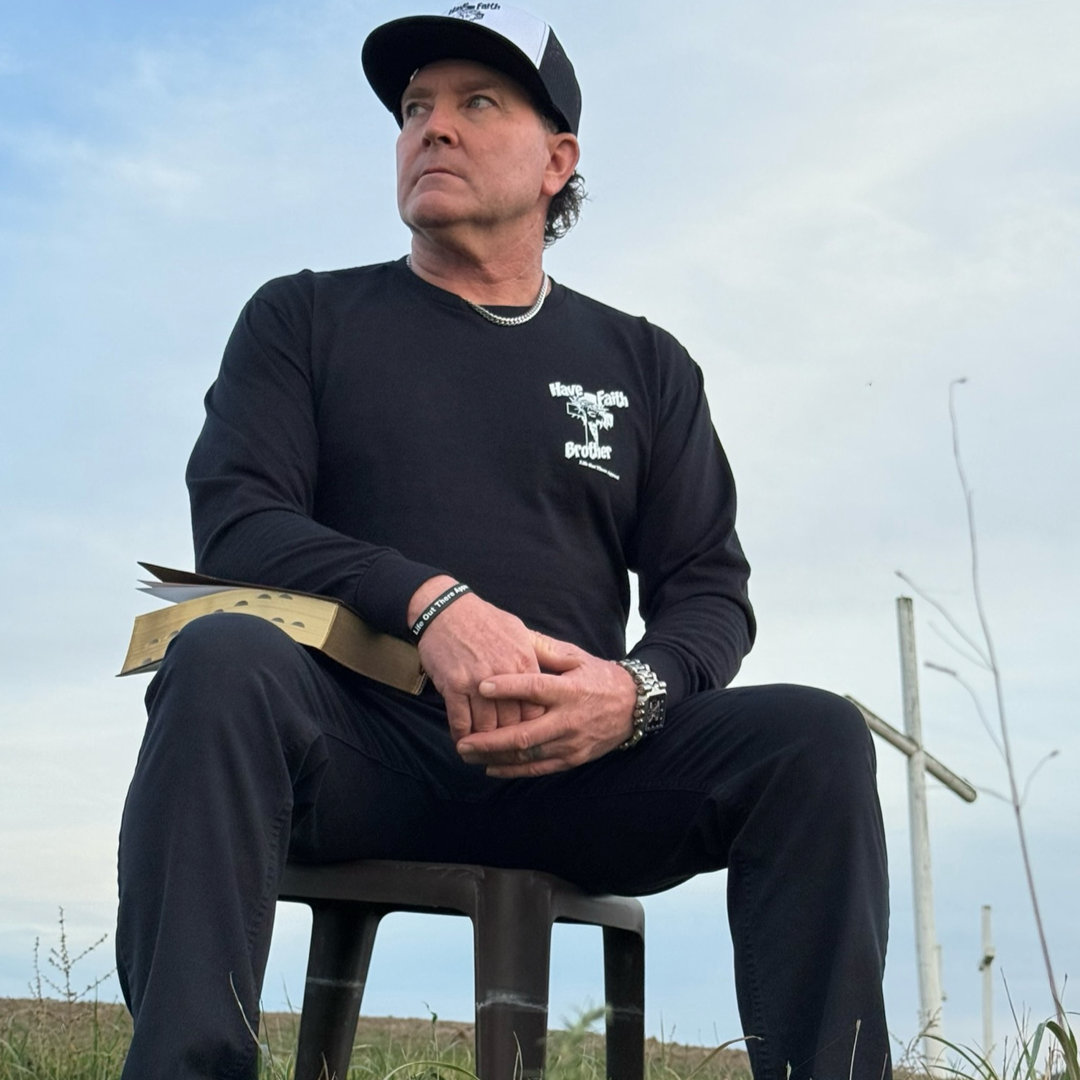 A man in the Life Out There Apparel Unisex "Have Faith Brother" Black Long Sleeve Tee sits outdoors holding a book, wearing black pants and a cap. He looks to the side seriously as a wooden cross stands in the background.