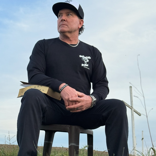 A man in the Life Out There Apparel Unisex "Have Faith Brother" Black Long Sleeve Tee sits outdoors holding a book, wearing black pants and a cap. He looks to the side seriously as a wooden cross stands in the background.