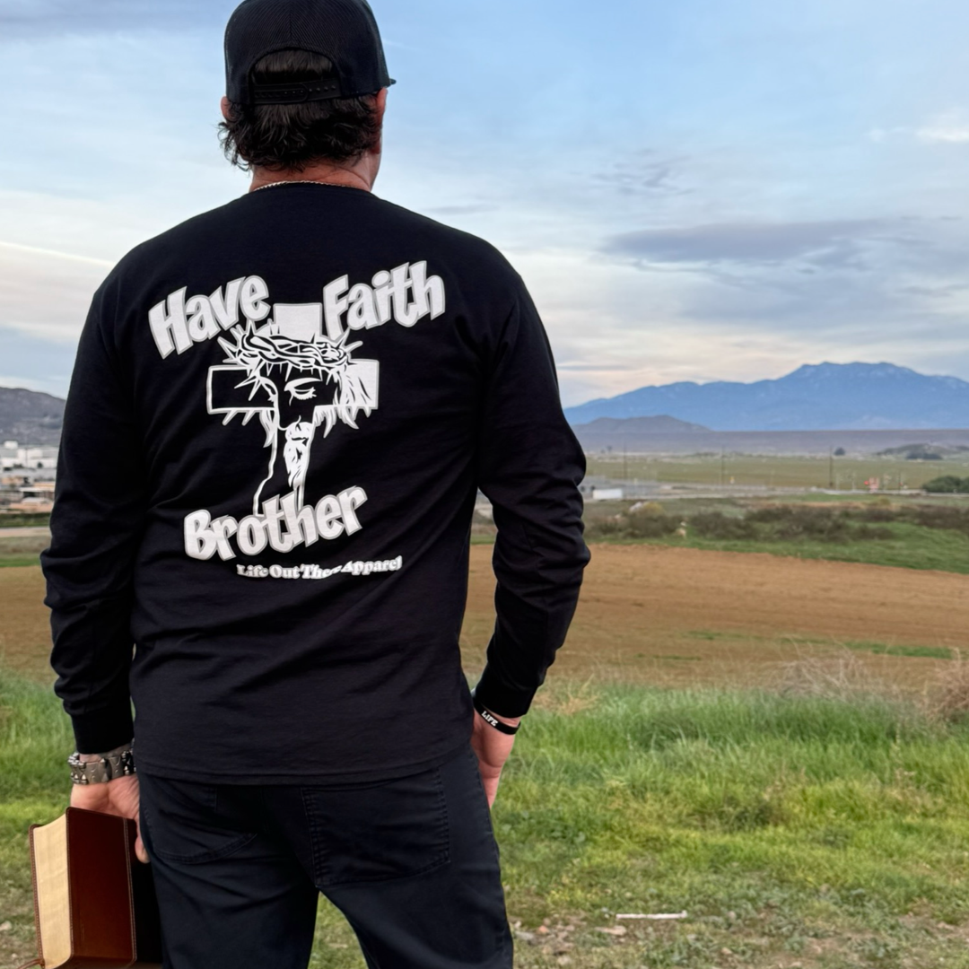 A person stands on grass, facing away, wearing the Life Out There Apparel Unisex "Have Faith Brother" Long Sleeve Tee in black—Christian faith clothing featuring a cross and crown of thorns, with fields and mountains in the distance.