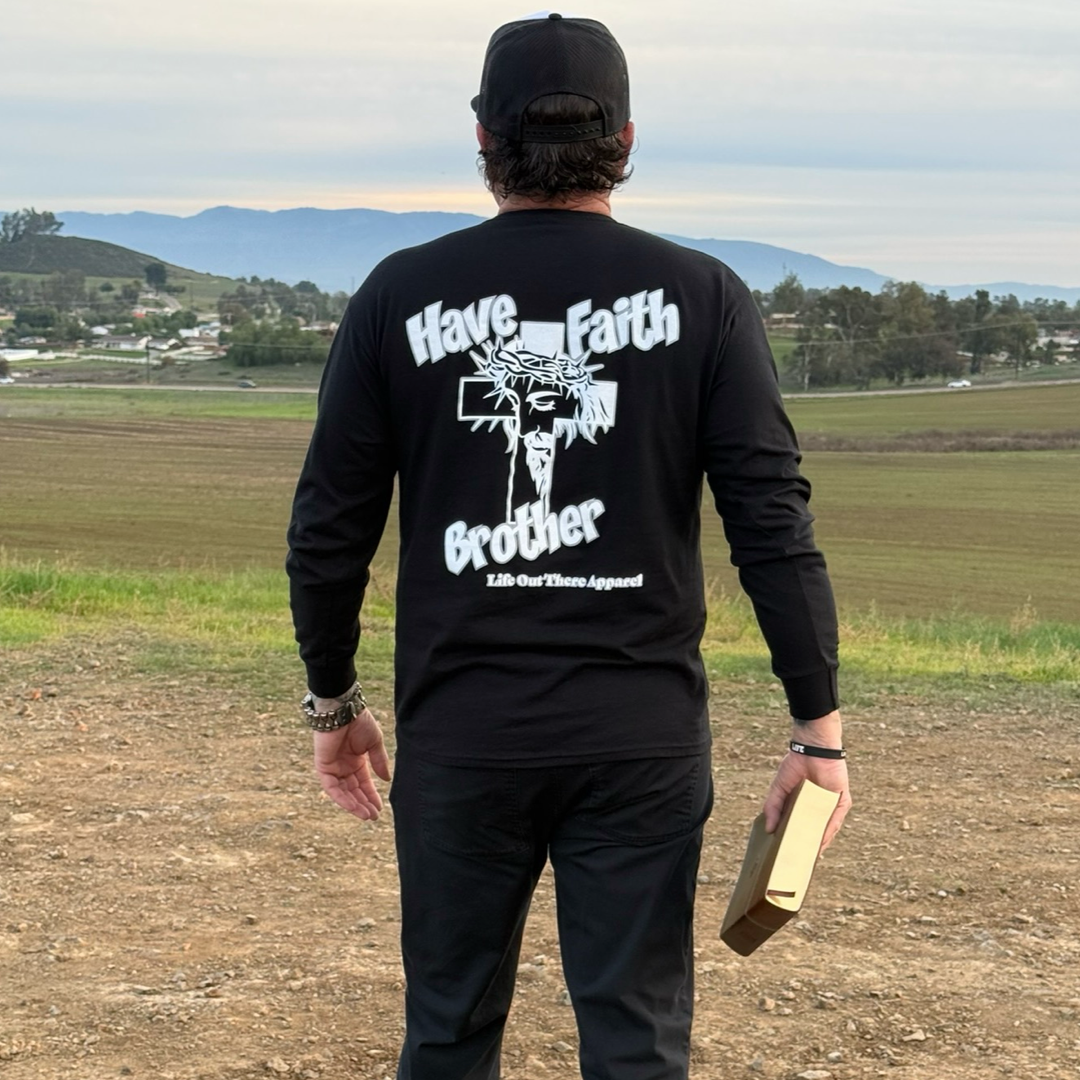 A man stands outdoors on a dirt path wearing the Unisex - Have Faith Brother - Long Sleeve Tee in black from Life Out There Apparel. He holds a book and wears a black cap, with fields and hills behind him.