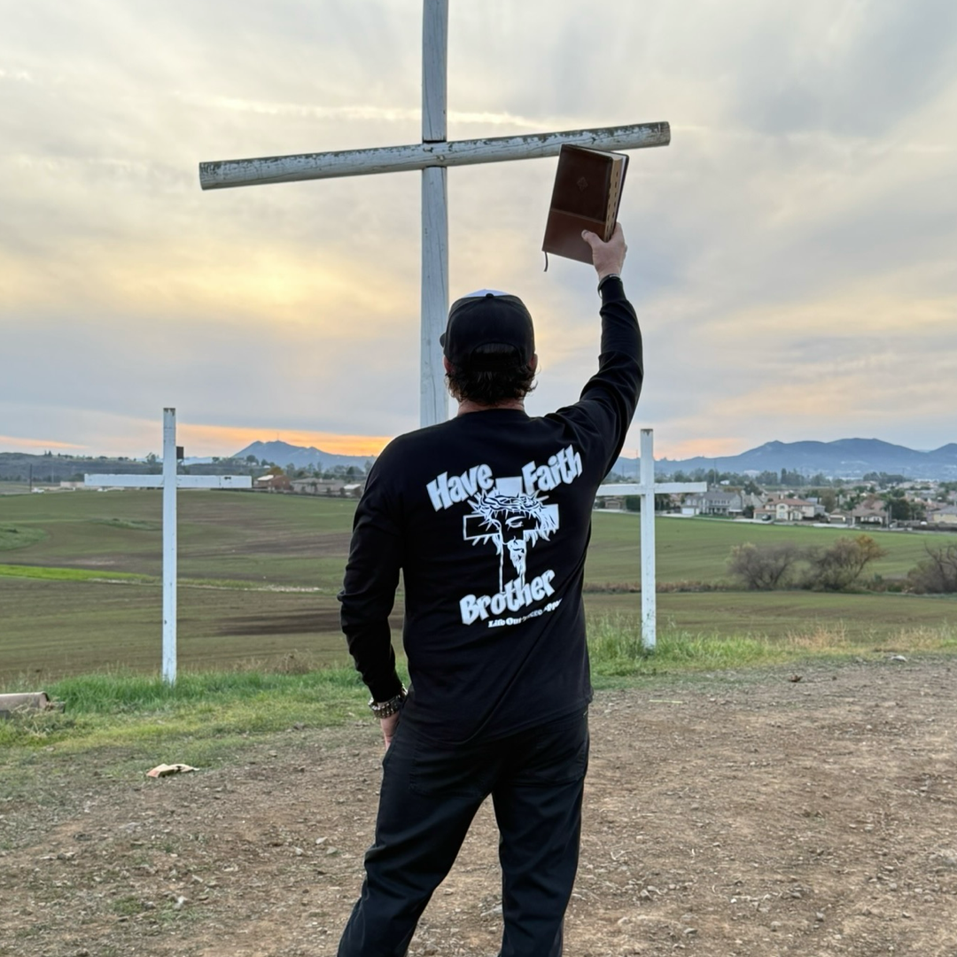 A person stands outdoors facing a cross, holding a Bible and wearing the Life Out There Apparel Unisex "Have Faith Brother" Long Sleeve Tee in black, overlooking fields and hills beneath a cloudy sky.