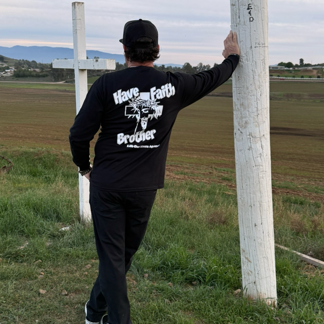 A person outdoors, wearing the Life Out There Apparel Unisex "Have Faith Brother" Long Sleeve Tee in black, leans on a large white cross. The shirt's back displays "Have Faith Brother," with fields and hills in the background.