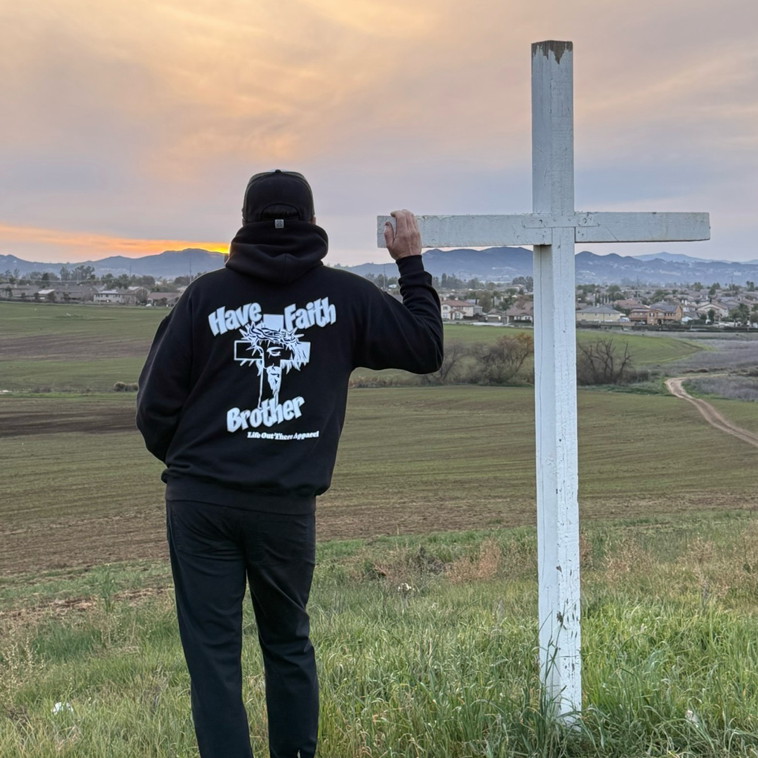 Wearing the Unisex - Have Faith Brother Hoodie - Black by Life Out There Apparel, a person stands on grass with a white cross at sunset, embodying Christian faith and inspiration among fields and distant mountains.