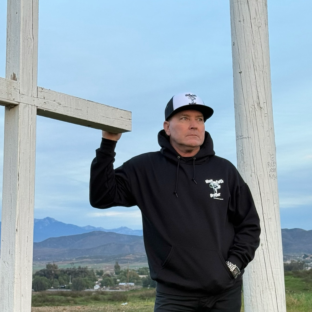 A man in a black Unisex - Have Faith Brother Hoodie by Life Out There Apparel leans against a large white wooden structure outdoors, with mountains and a cloudy sky in the background.