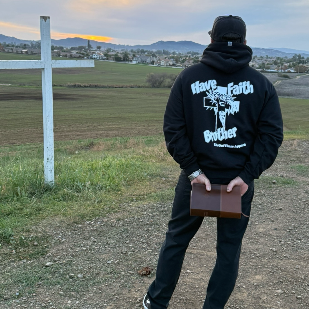 At sunset near a white cross, a person faces a field and distant town, wearing the black "Have Faith Brother" unisex hoodie by Life Out There Apparel and holding a closed book behind their back.