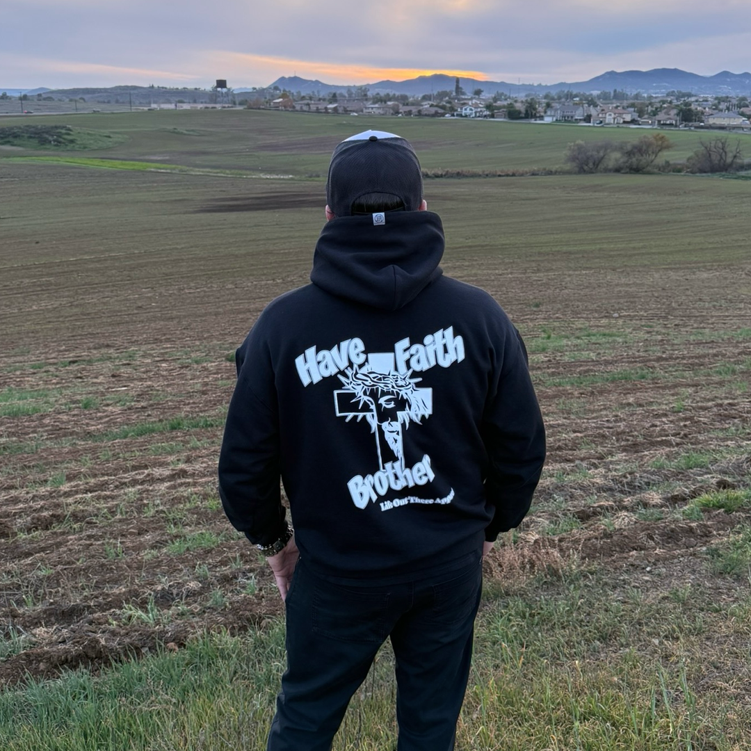A person stands in a field at sunset, wearing the black Unisex - Have Faith Brother Hoodie by Life Out There Apparel, featuring a cross and "Have Faith Brother" on the back. Houses, hills, and a cloudy sky complete the peaceful scene.