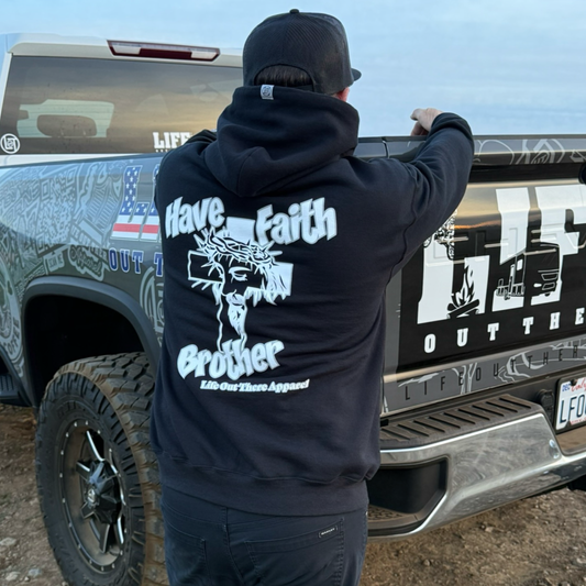 Wearing the Unisex - Have Faith Brother Hoodie - Black by Life Out There Apparel, a person stands by a decorated pickup truck with "LIFE OUTF" on the side, blending Christian faith and casual style.