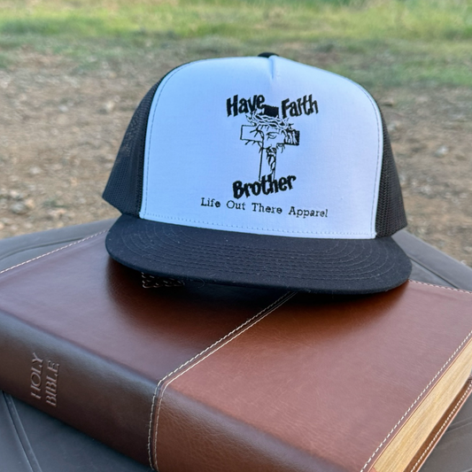 The Have Faith Brother (Embroidered) Flat Bill SnapBack Trucker Hat in white and black by Life Out There Apparel rests on a closed Holy Bible outdoors, with grass and gravel visible in the background.