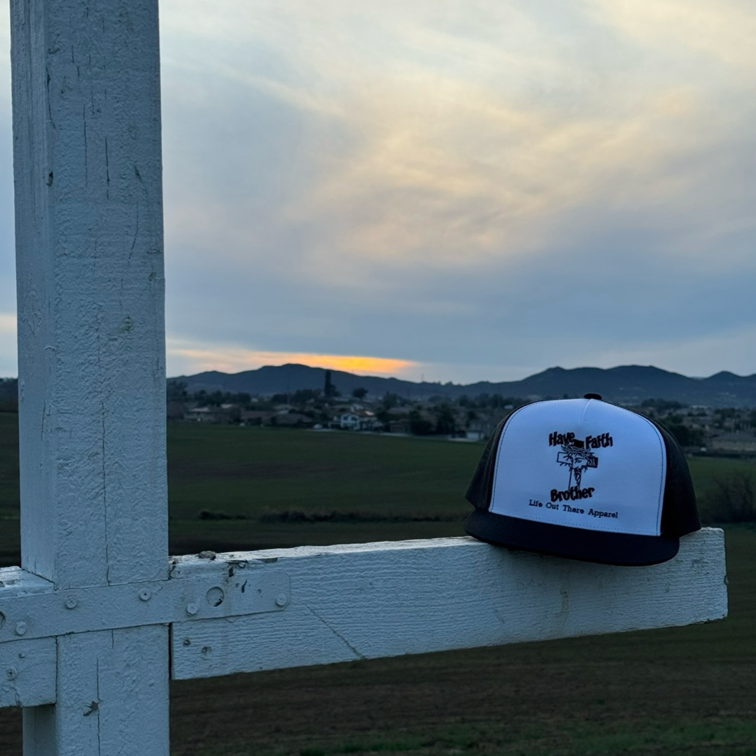 A Life Out There Apparel Have Faith Brother (Embroidered) Flat Bill Snapback Trucker Hat in White/Black sits on a white wooden fence at sunset, overlooking fields and hills beneath a cloudy sky.