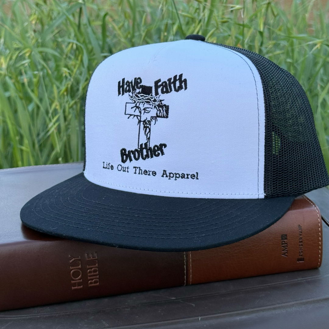 The Have Faith Brother (Embroidered) Flat Bill SnapBack Trucker Hat in white/black by Life Out There Apparel rests on a closed Holy Bible outdoors, green grass softly blurred in the background.